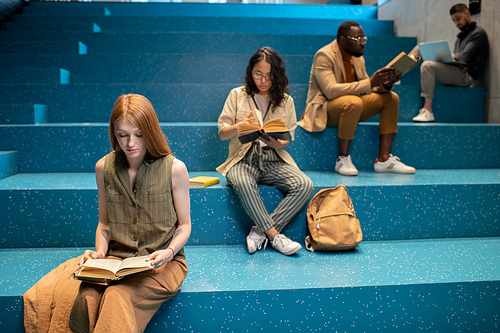 Group of intercultural diligent students sitting on blue staircase and reading