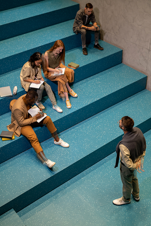 Group of diligent students making notes in copybooks while sitting in front of coach