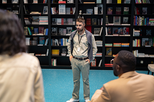 Strict young teacher or coach standing in front of intercultural students in library or bookstore