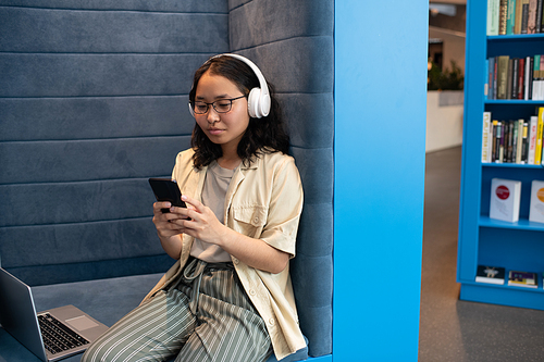 Young female in headphones scrolling in smartphone while spending time in bookstore