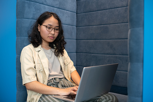 Young Asian woman in eyeglasses and casualwear networking in front of laptop