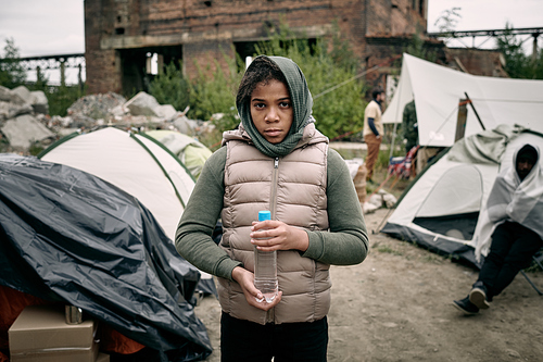 Cute girl with bottle of water standing in front of camera against tents of refugee settlement