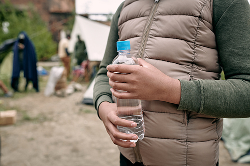 Close-up of unrecognizable black girl in warm vest standing with bottle of water against migrant camp