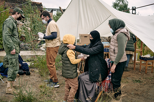 Young Muslim woman in protective mask getting her daughter dressed with volunteers on background