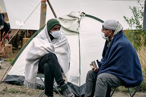 Middle eastern male migrants in masks sitting covered with blankets at tent and talking