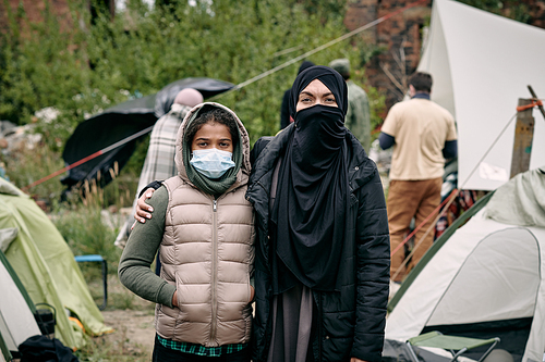 Young woman in hijab and her daughter standing against tent camp for migrants