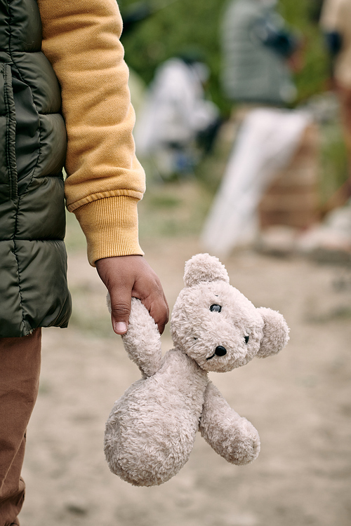 Close-up of unrecognizable refugee child in sweater and vest walking with plush bear through ruins