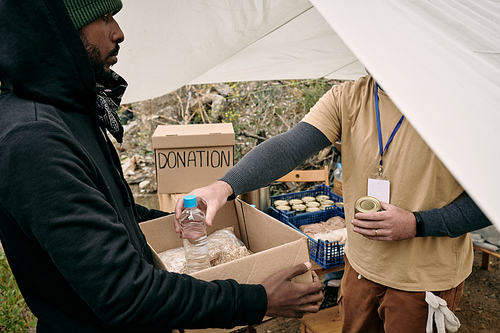 Young refugee man holding box while volunteer putting there food and water for the family of migrants