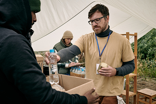 Young bearded volunteer in glasses holding tin can and giving bottle of water to homeless man with box