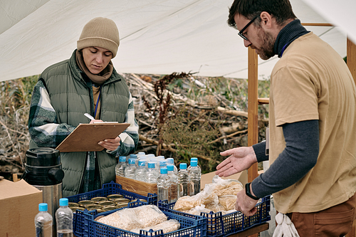 Female volunteer making notes in clipboard while distributing food with assistant to refugees