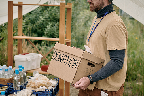 Bearded volunteer with badge carrying cardboard box with donation to table with goods for homeless