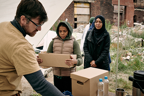 Young bearded social employee in glasses giving food and water to black refugee girl with open box outdoors