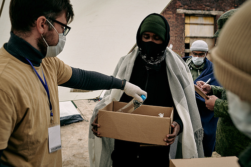 Social Worker in mask and glasses putting bottle of water into box held by middle-eastern refugee with plaid on shoulders