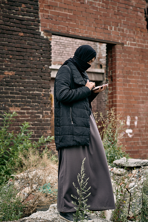 Middle-eastern refugee woman in hijab and long dress standing against abandoned building and using phone