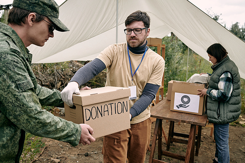 Serious young bearded social worker with badge passing donation box to soldier outdoors
