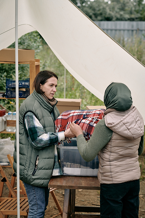 Young female volunteer offering warm woolen scarf to Muslim woman living in refugee camp