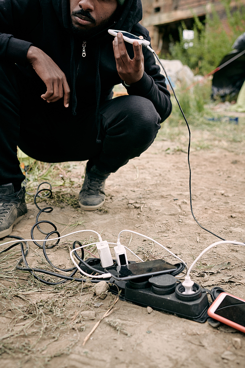 Close-up of black refugee man crouching at socket outlet and listening to audio message on phone while charging it outdoors