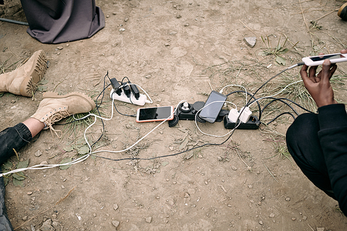 Hand of young refugee holding smartphone on charge while sitting by other gadgets, plugs and chargers