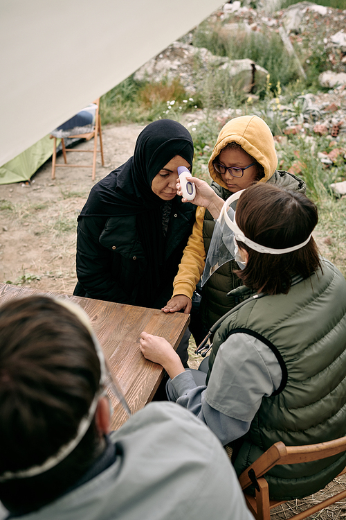 Volunteering doctor measuring body temperature of young female in hijab on territory of refugee camp