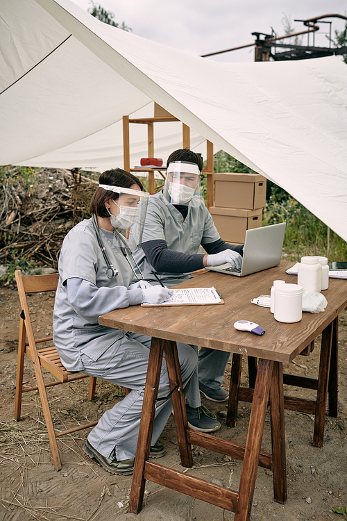 Team of doctors in safety face shields sitting at table in tent and working with medical cards of refugees at remote area