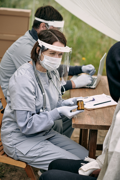 Female nurse in plastic screen mask and gloves sitting at table outdoors and providing medication to migrants
