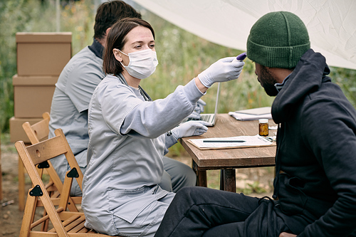Doctor in protective mask measuring body temperature of sick man from migration camp