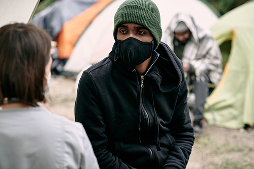 Sick young Black refugee man in facial mask and hat talking to nurse in migrant camp
