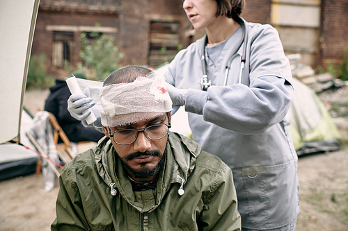 Young doctor in uniform putting bandage around wounded head of refugee man