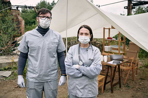 Portrait of young medical workers in masks and scrubs standing against tent with medicine in refugee camp