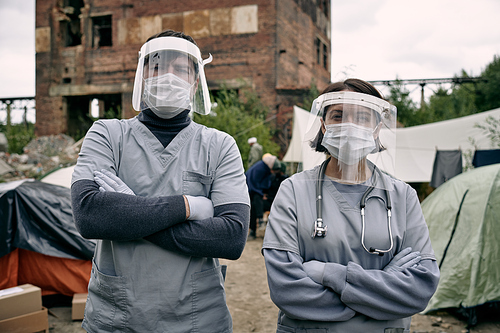 Two young clincians in protective workwear standing against tents of migrants in refugee camp