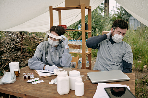 Exhausted ealthcare workers in masks sitting at table with pills under tent roof while working with refugees in camp