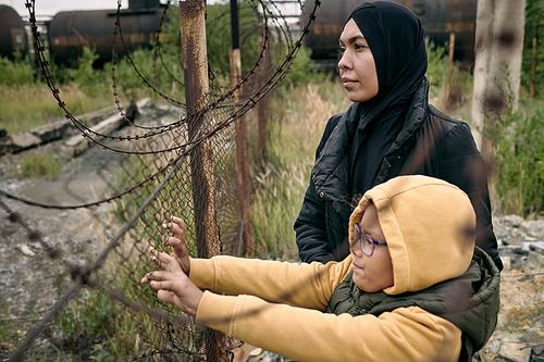 Hopeful Muslim woman in hijab standing with daughter at barbed wire and looking into distance