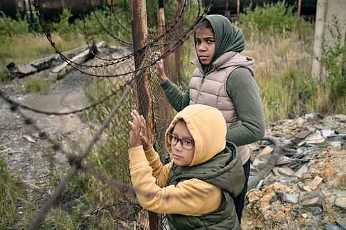 Two little girls in hoodies and jackets standing by net dividing zone of refugee camp from other territory