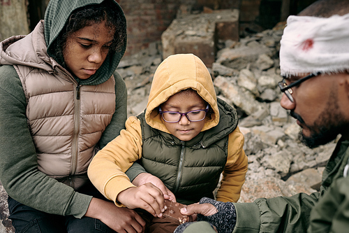 Sad middle-eastern refugee family sitting on ruins of building and sharing chocolate