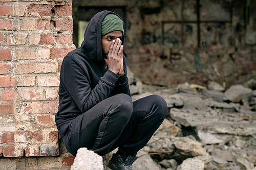 Distraught young Black refugee man in black outfit crouching at ruined building and covering face with hands
