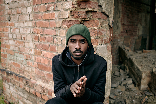 Young African man in hoodie and cap sitting by brick wall of ruined building
