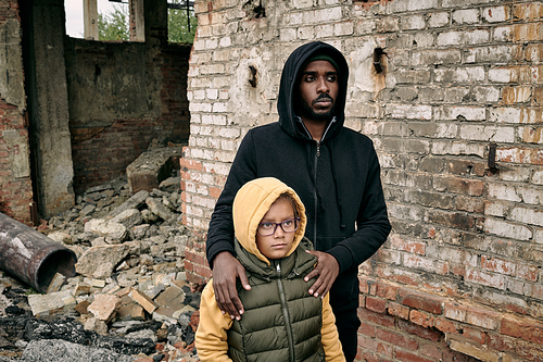Sorrowful young Black man in hoodie embracing little daughter around shoulders while standign with her against ruined house