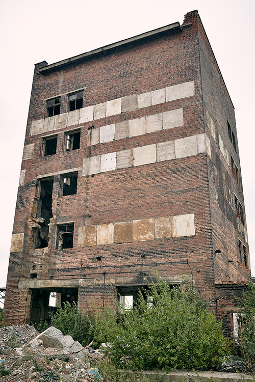 Gloomy abandoned brick building with broken windows and bushes, stones around first floor