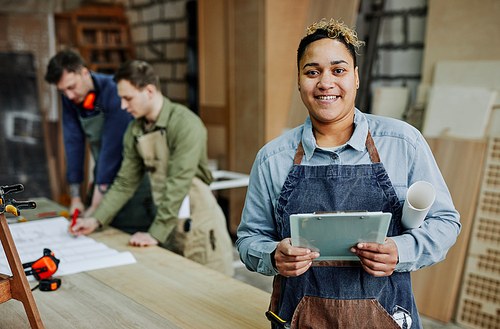 Waist up portrait of smiling female carpenter holding clipboard and looking at camera while posing in artisan workshop, copy space