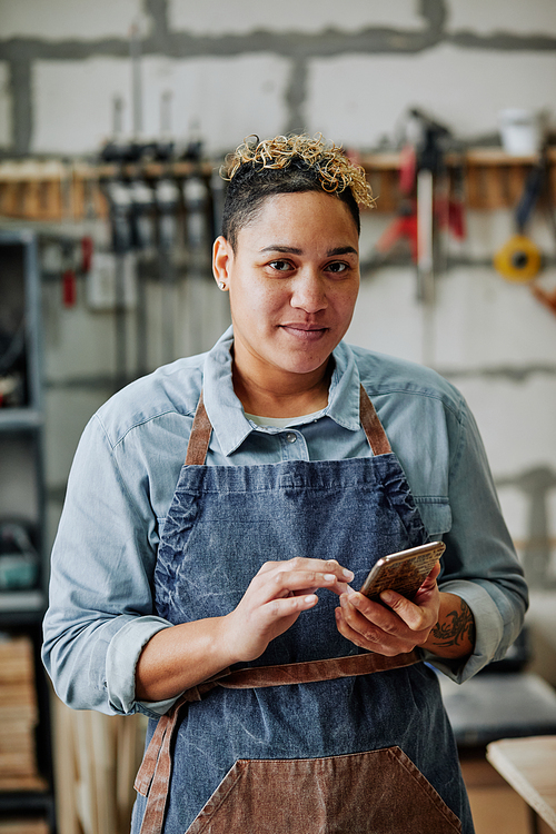 Vertical waist up portrait of female artisan using smartphone and smiling at camera while standing in workshop with tools in background