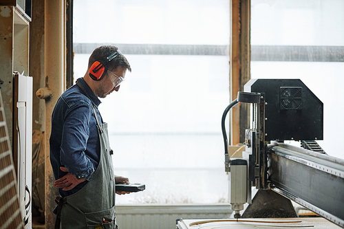 Side view portrait of male carpenter operating CNC cutting machine in automated production workshop