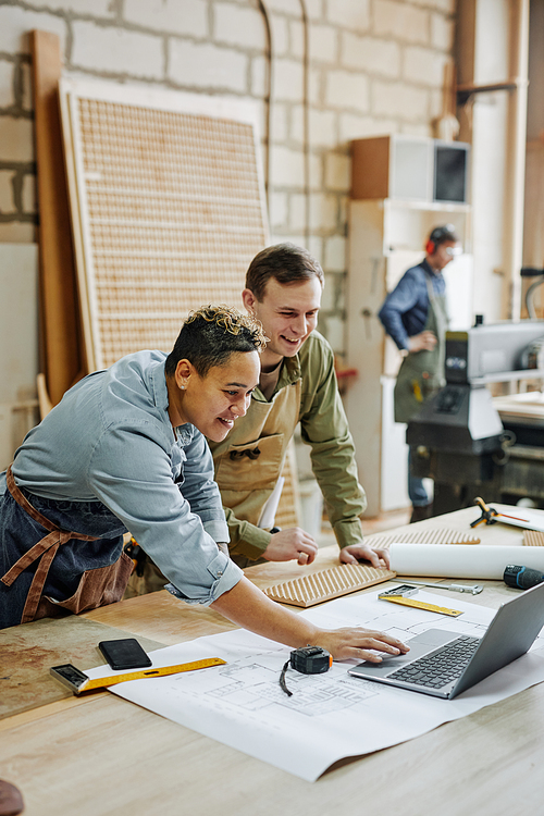 Vertical warm toned shot of two modern carpenters using laptop in workshop and discussing designs