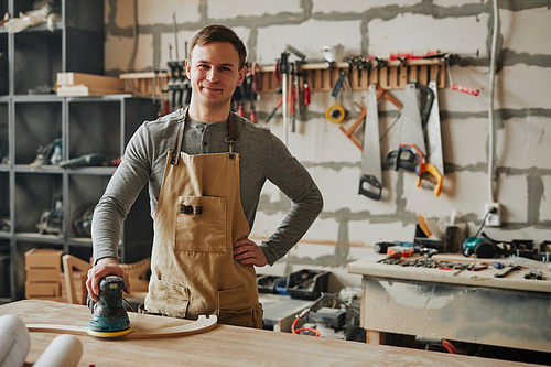 Warm toned shot of young carpenter smiling at camera while building furniture in workshop, copy space