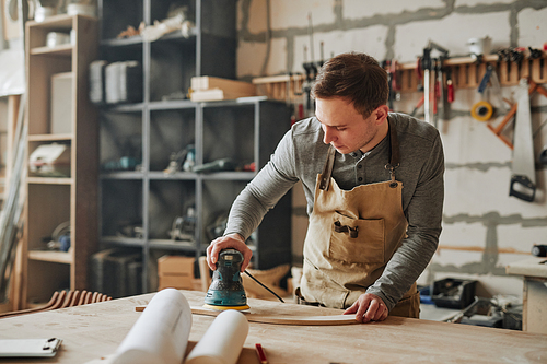 Warm toned portrait of young carpenter sanding wood and building handmade furniture piece in workshop, copy space