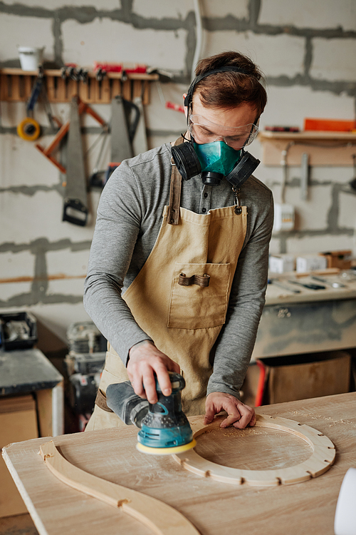 Warm toned portrait of young carpenter in protective gear working with wood while building handicraft furniture in workshop