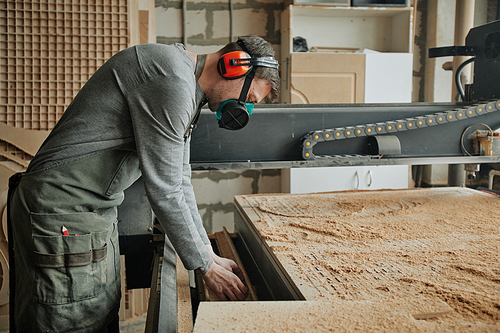 Side view portrait of male worker wearing full protective gear while operating machines at woodwork production