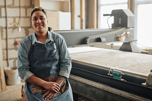 Portrait of smiling female worker looking at camera while posing by woodcutting machine in workshop, copy space