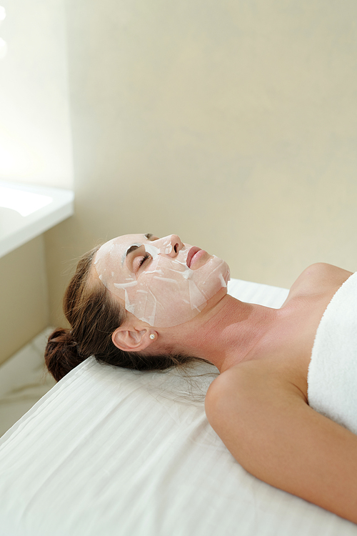 Young relaxed female with sheet mask on face enjoying skincare procedure in luxurious spa salon