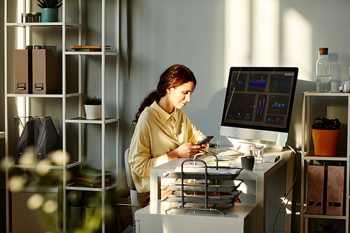 Young adult Caucasian women wearing casual shirt sitting at desk in modern office scrolling news feed in social net on smartphone