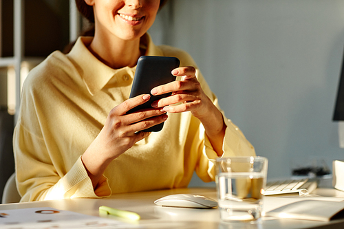 Medium section shot young adult woman wearing beige shirt sitting at desk in office room texting message on smartphone